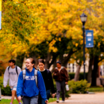 Group of students walking on a college campus with fall foliage and banners in the background, carrying backpacks.