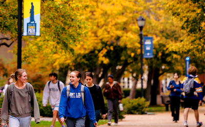 Group of students walking on a college campus with fall foliage and banners in the background, carrying backpacks.