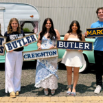 Five young adults posing outdoors in a parking lot, each holding college name signs: Saint Mary’s, Butler, Creighton, Butler, and Marquette, with a vintage trailer and car in the background.