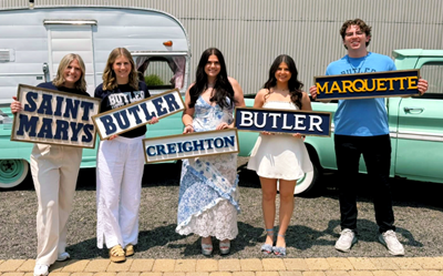Five young adults posing outdoors in a parking lot, each holding college name signs: Saint Mary’s, Butler, Creighton, Butler, and Marquette, with a vintage trailer and car in the background.