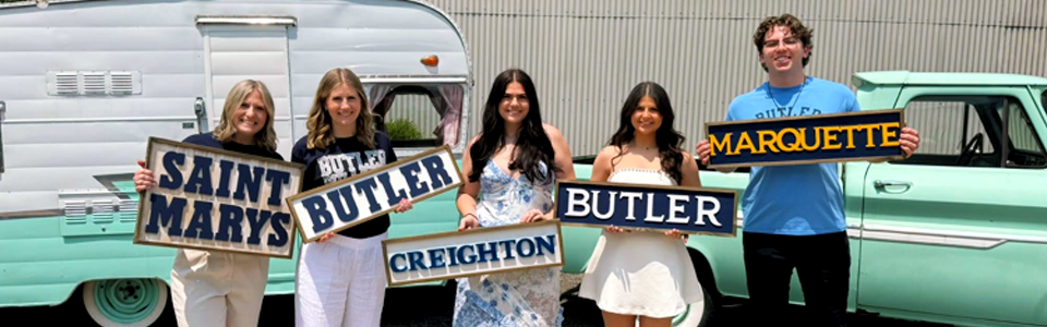 Five friends pose in front of a vintage camper and mint-green car, holding signs for Saint Mary’s, Butler, Creighton, Butler, and Marquette.