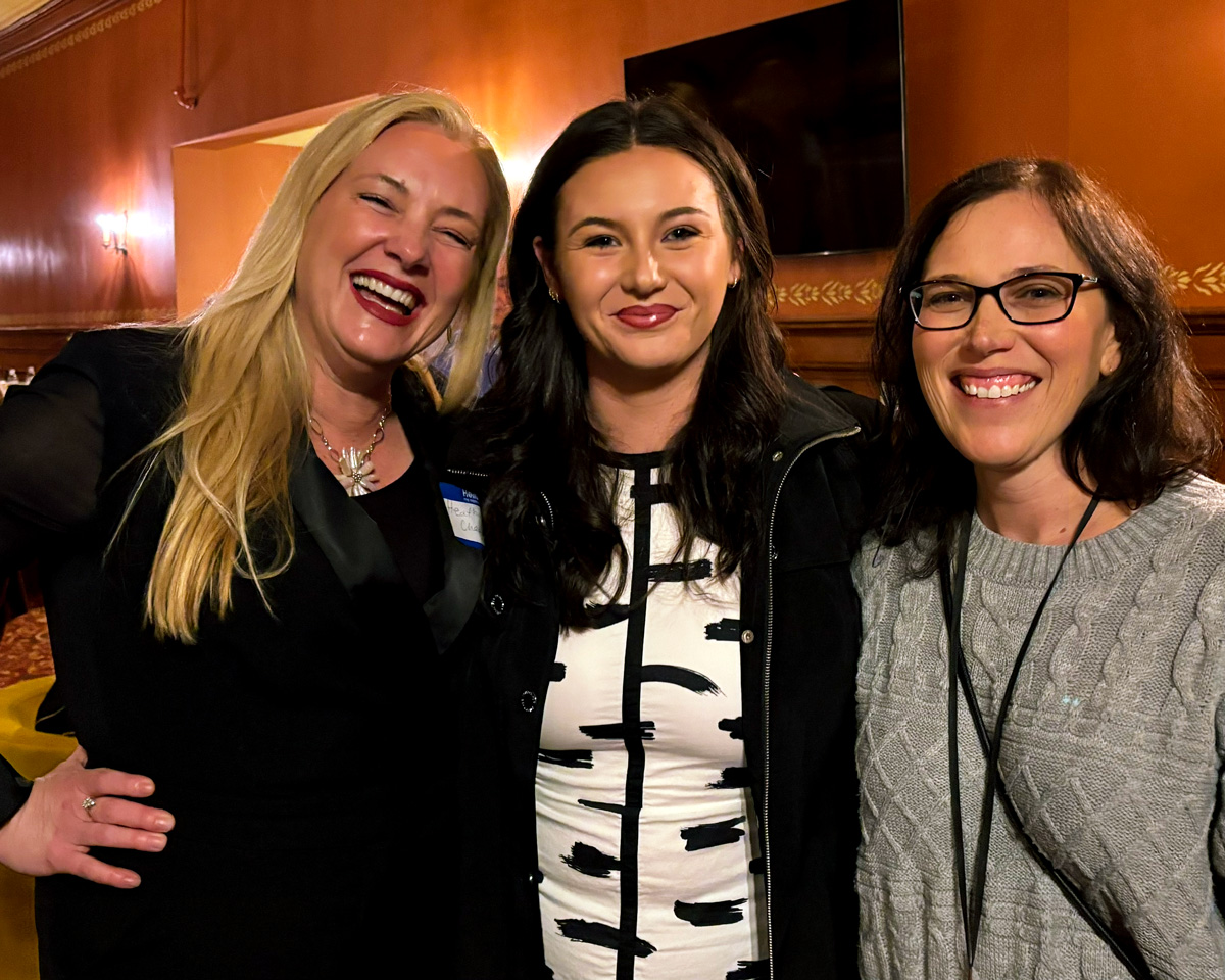 Three women smiling and posing together at an indoor event.