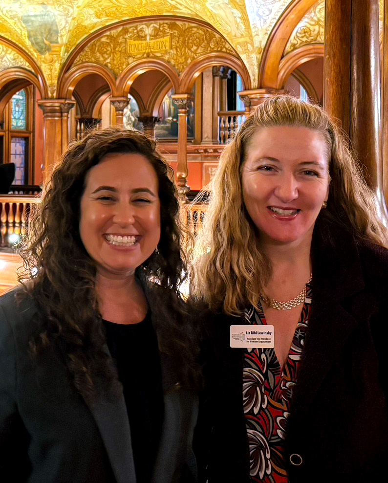 Two women in business attire smiling at the camera inside a gold-decorated, arch-filled hall; one wears a name badge on her blazer.