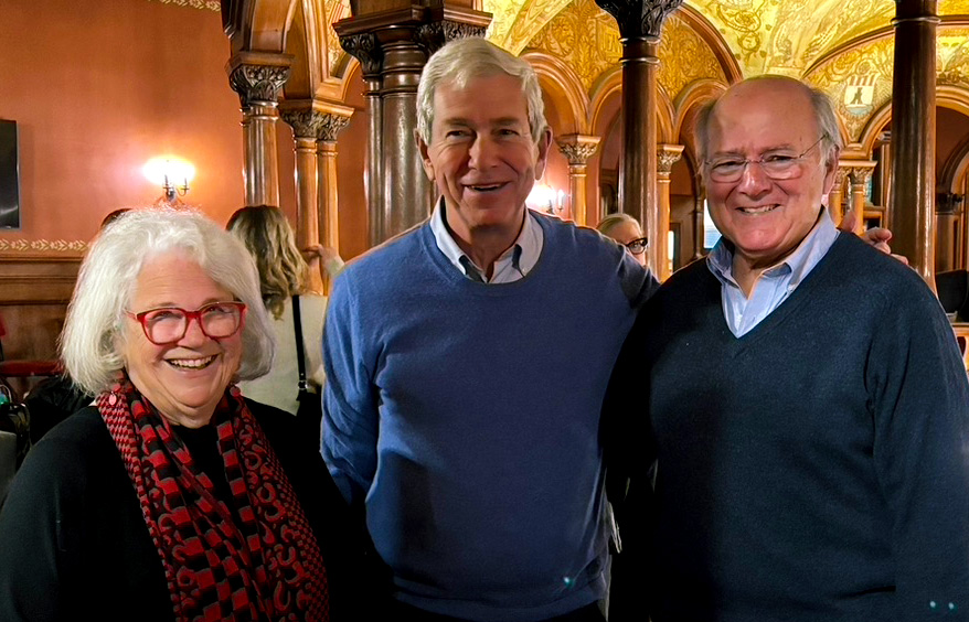 Three adults posing together and smiling in a wood-paneled, ornately decorated room with arches and columns.