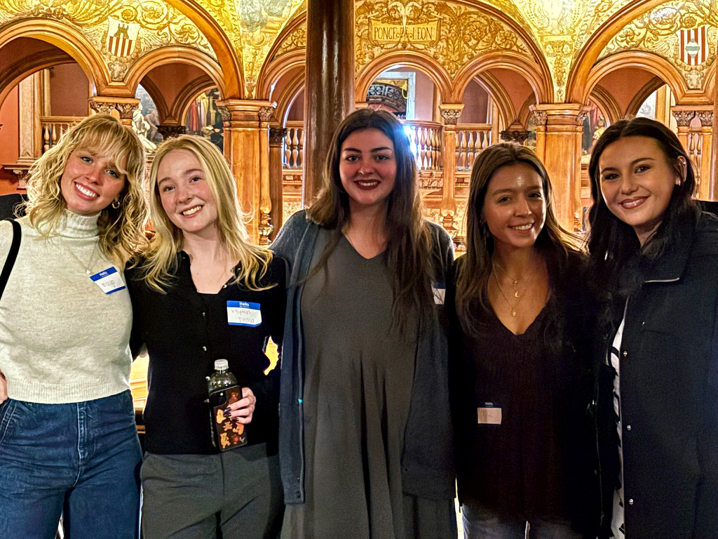 Five smiling young women stand side by side in a gilded, arched hall at an event, wearing name tags with one holding a bottle of tea/soda nearby.