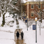 Winter campus courtyard: two people walk down a cleared brick path between snow-covered trees and red brick buildings.