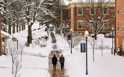 Winter campus courtyard: two people walk down a cleared brick path between snow-covered trees and red brick buildings.