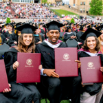 Group of diverse graduates in caps and gowns seated on a field, each holding a maroon diploma cover and smiling at the camera at a ceremony.