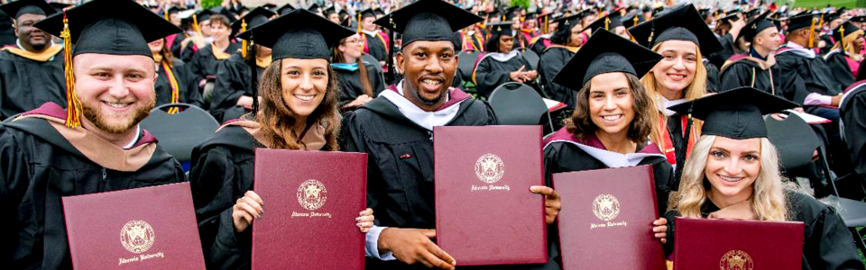 Group of graduates in caps and gowns posing with diplomas at a commencement ceremony with smiling faces in the front row.